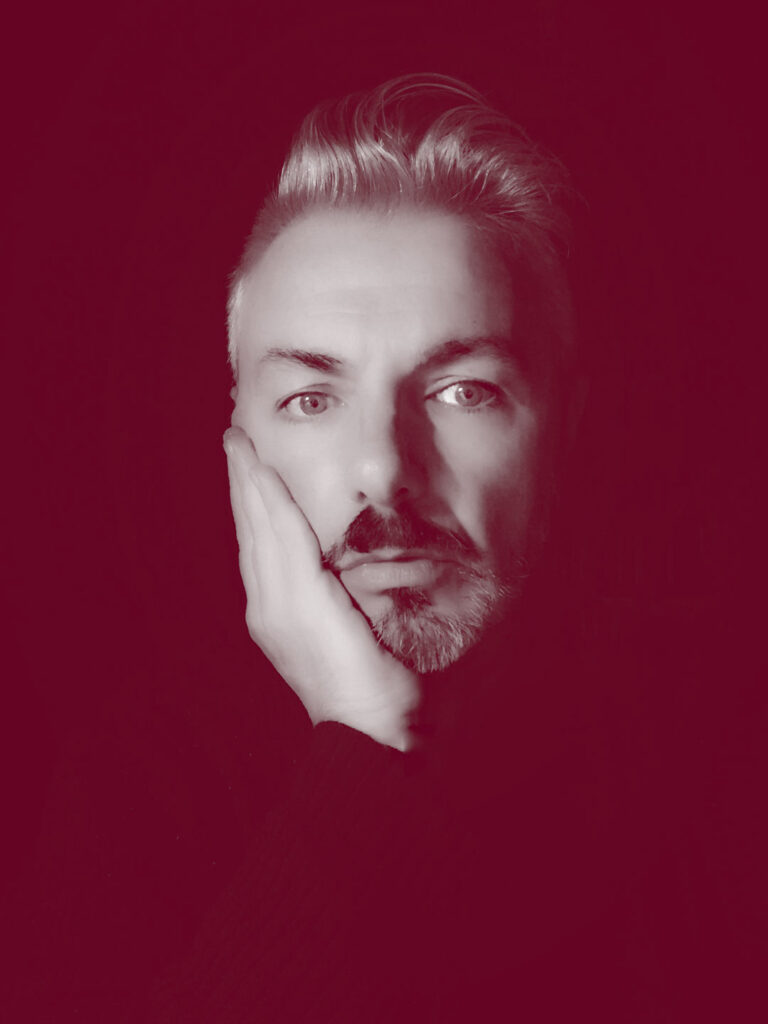 In primo piano il viso di Alessandro Tampieri, un uomo con i capelli e barba brizzolati. La mano destra è appoggiata sulla guancia destra.