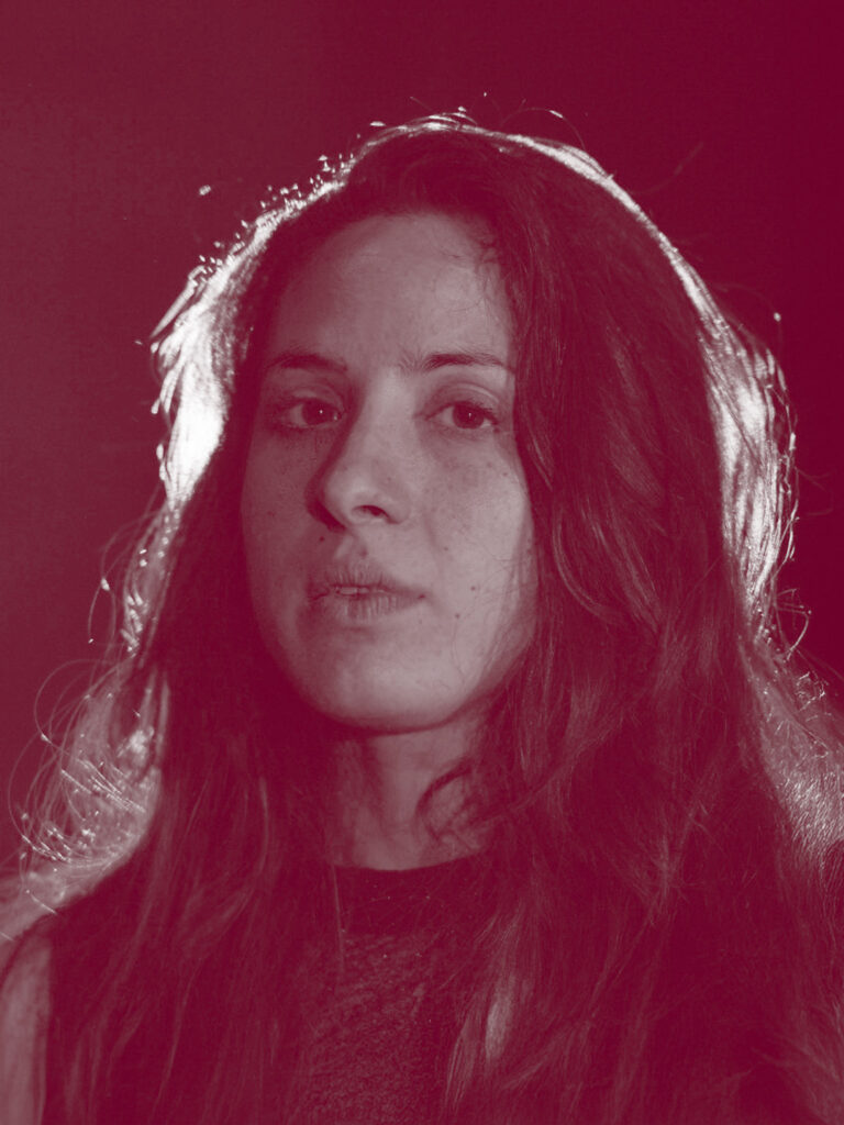 In primo piano Camilla Guarino, una donna con i capelli scuri e lunghi, tenuti sciolti che cadono sulle spalle. Sulle labbra un lieve sorriso.