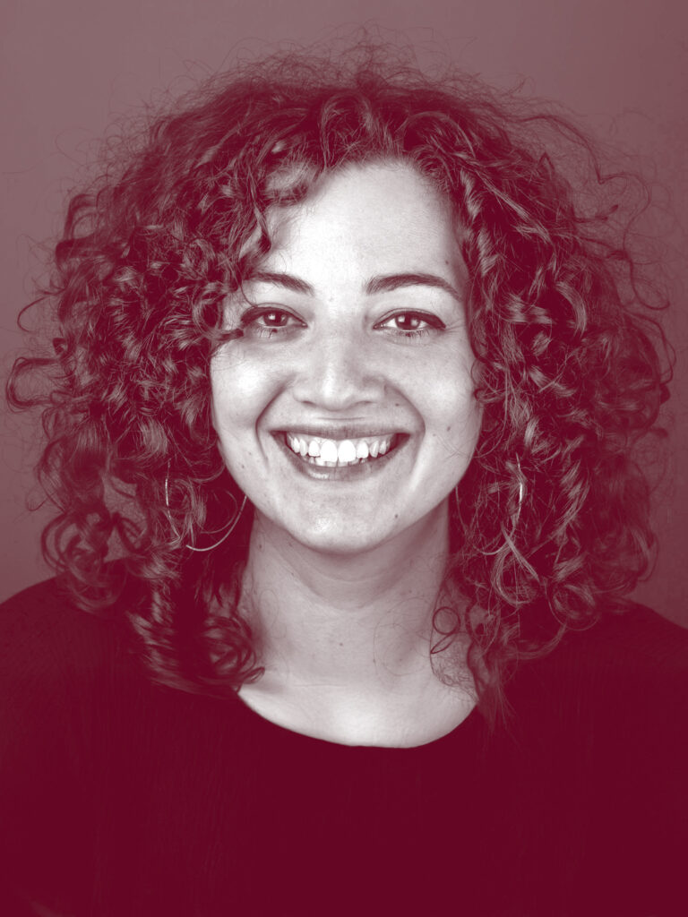 In primo piano Cristina Cavallo, una persona sorridente con i capelli ricci lunghi fino alle spalle che le circondano il viso sorridente, i suoi occhi sono truccati con eyeliner e due piccoli pallini sulla palpebra inferiore. Indossa una maglietta nera con scollo rotondo.