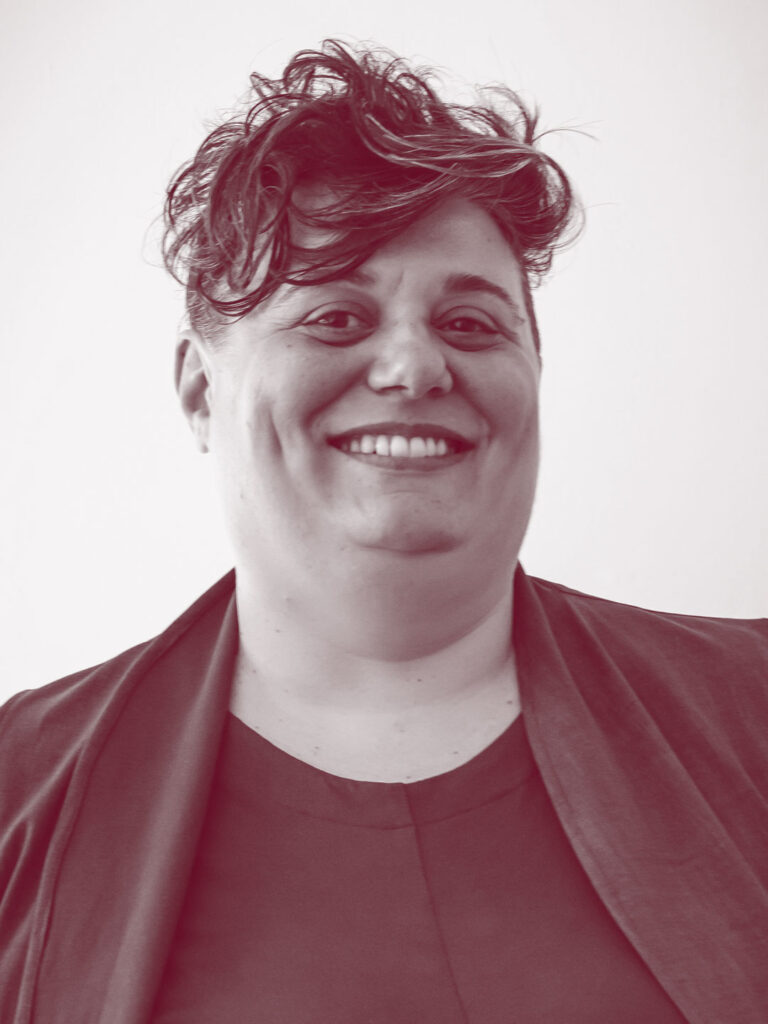 In primo piano Elisa Coco, una persona con i capelli corti e mossi e un grande sorriso. Indossa una maglietta e una giacca nera.