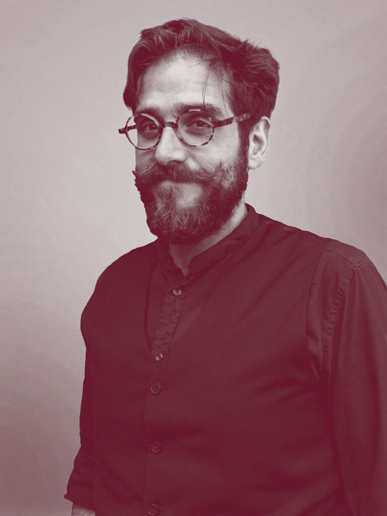 La foto è in bianco e nero. Mezzo busto di Luca Falbo, una persona con i capelli corti castani, occhi marroni, occhiali e barba. Indossa una camicia e un gilet blu. Foto di Carlotta Stracchi Villa.