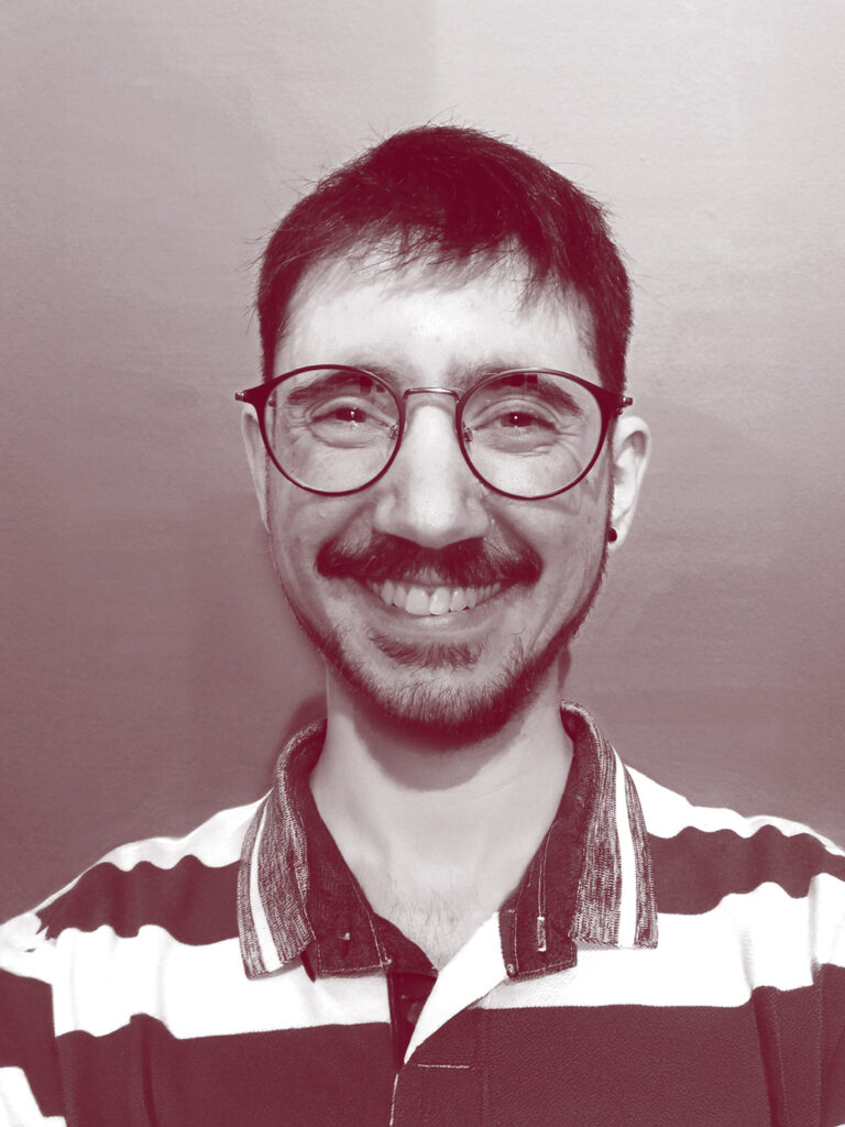 In primo piano il viso di Ralph Pacinotti, una persona con gli occhiali da vista, i capelli e la barba scuri. Indossa una polo a righe con le maniche corte e sorride.