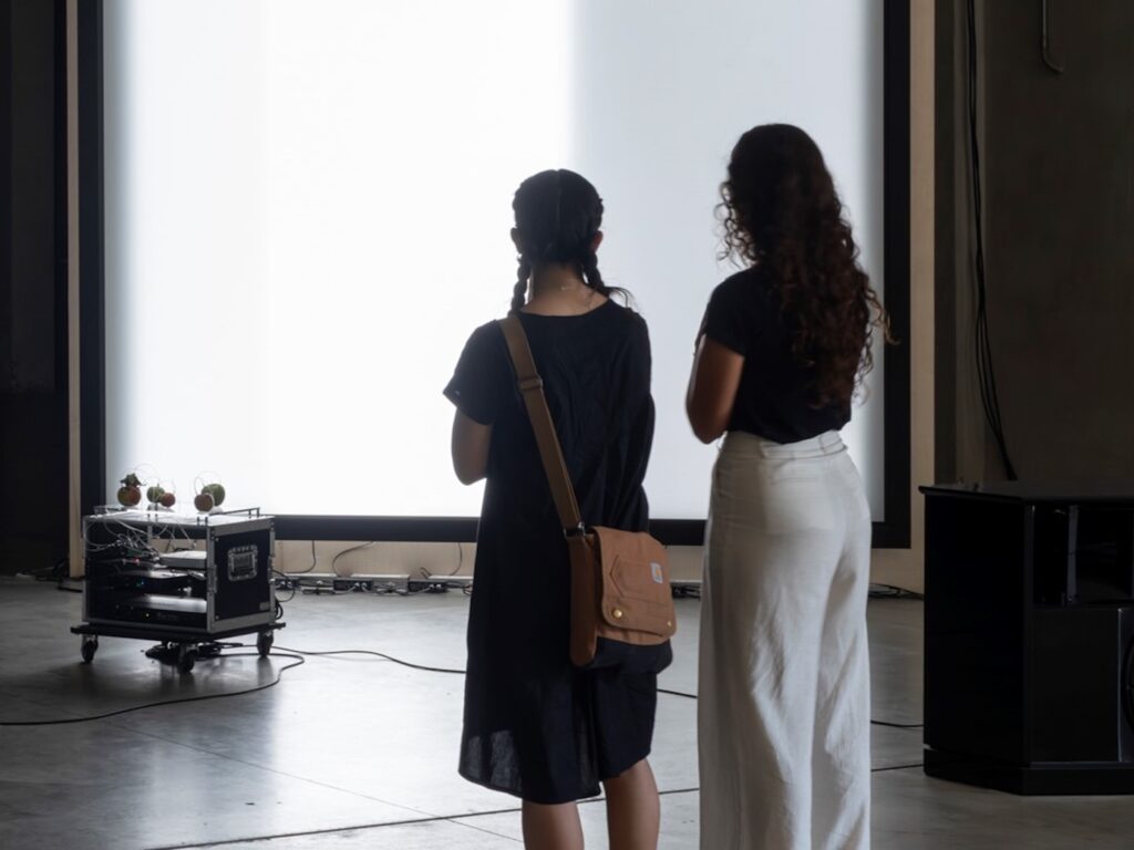 Veduta dell'allestimento della mostra. Due ragazze di spalle osservano l'opera posta sullo sfondo dell'immagine.