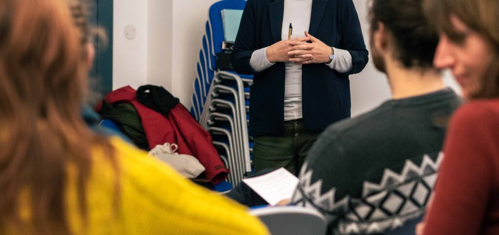 Foto scattata durante un incontro di formazione. Di spalle, le persone partecipanti; sullo sfondo, il busto dell'insegnante che si rivolge al pubblico.
