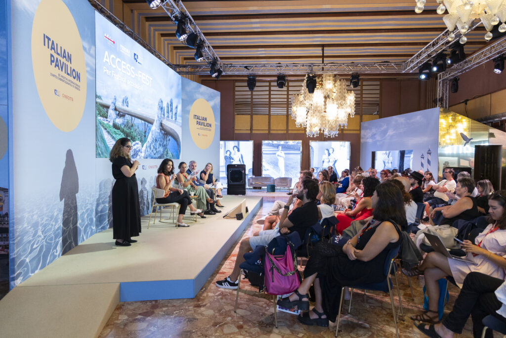 A destra, il pubblico; a sinistra, il palco su cui sono presenti l'interprete LIS, Valeria di Fedora e tre rappresentanti dell'Associazione festival italiani di cinema. La foto è stata scattata durante la presentazione della guida alla Mostra del Cinema di Venezia. Fotografia di Moris Puccio.